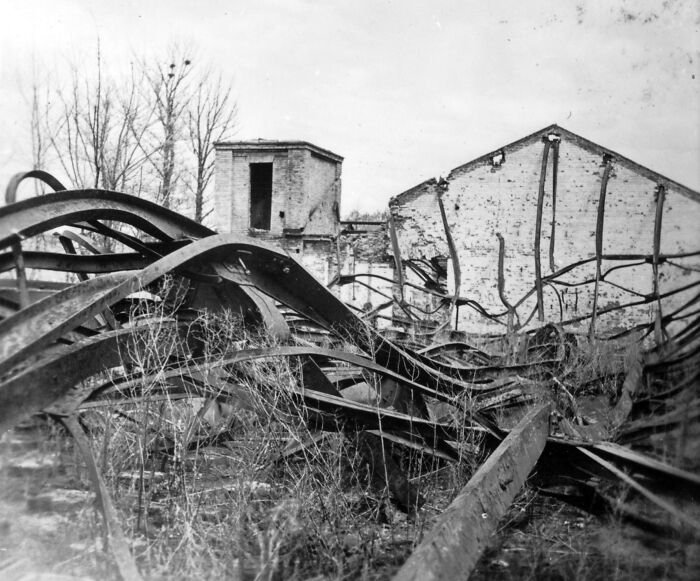 WWII photography showing twisted metal and a damaged brick building amidst barren trees.