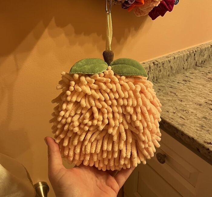 Hand holding a fuzzy peach-shaped find with green leaves, hanging in a bathroom setting.
