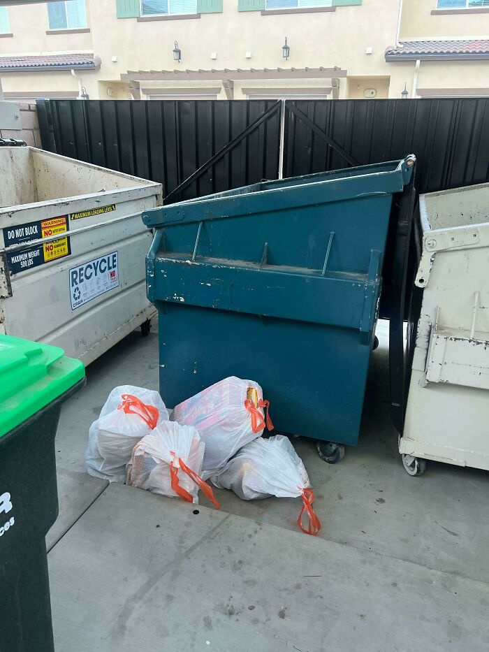 Trash bags with orange ties left beside dumpsters, illustrating lack of self-awareness in waste disposal.