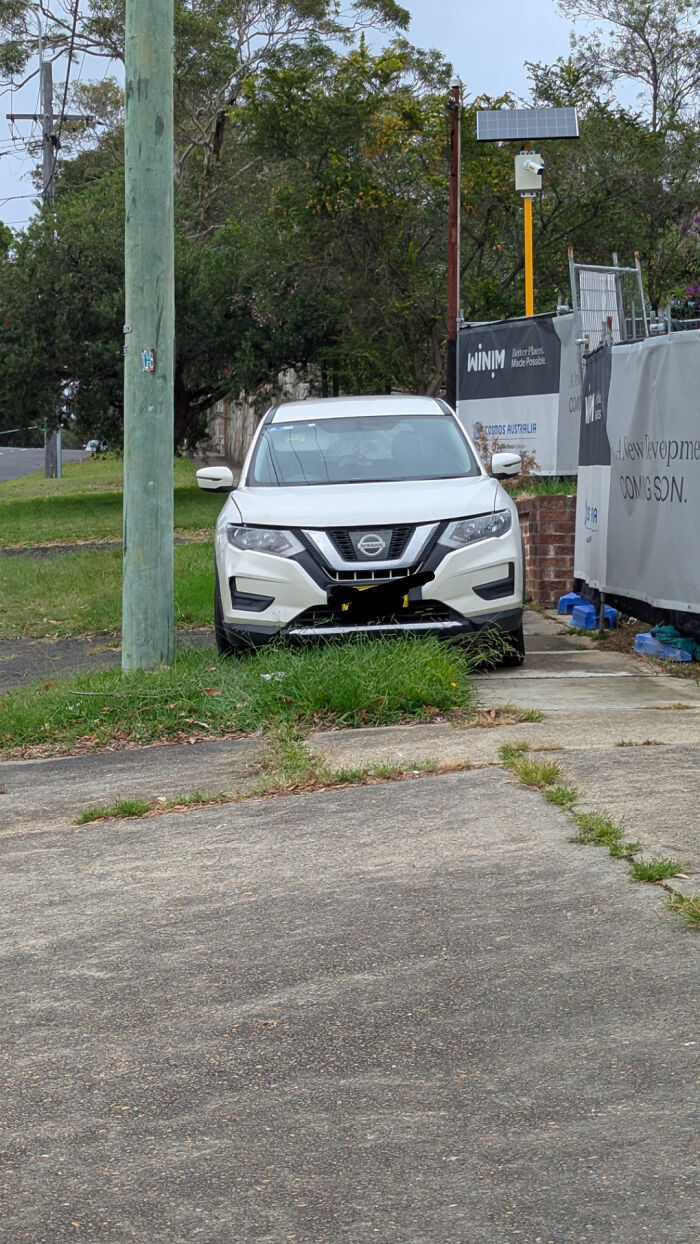 Car parked with no self-awareness, blocking sidewalk at a construction site with greenery around.