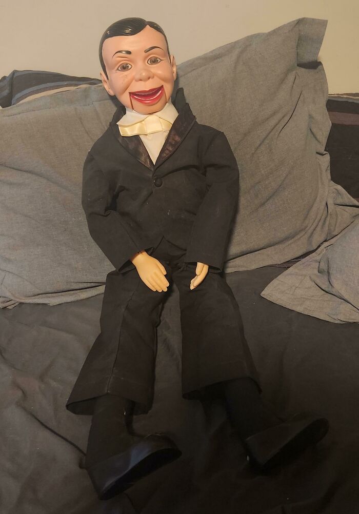 A ventriloquist dummy in a suit with a big smile sitting on a bed, representing funny elderly humor.
