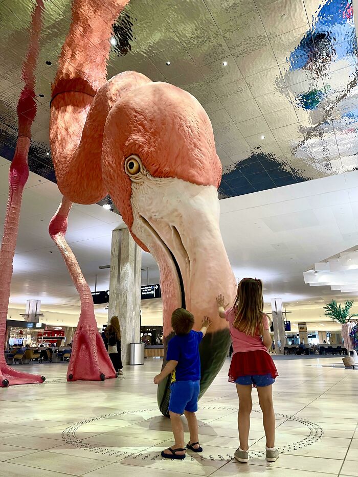 Children touching a massive flamingo sculpture in an airport, a photo that looks fake but is real.