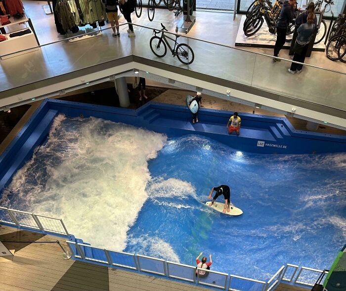 Indoor surfing wave looks fake, but it's a real pool setup in a store, with people watching from the upper level.