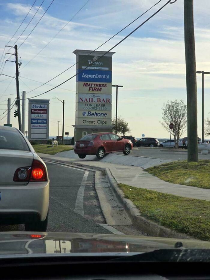 Red car awkwardly parked on a curb in front of shopping center signs, highlighting lack of self-awareness.