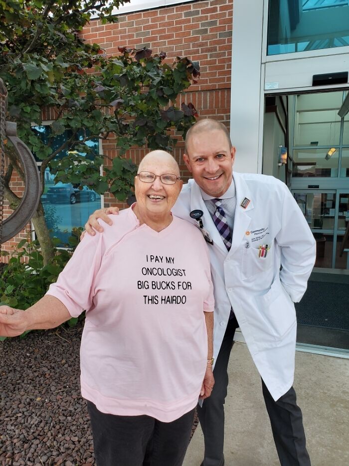 Elderly person with humorous shirt about hairdo, posing with doctor outside a building.