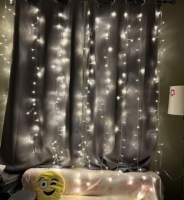 Fairy lights hanging on curtains above a couch with emoji pillow, evoking a nostalgic millennial vibe.