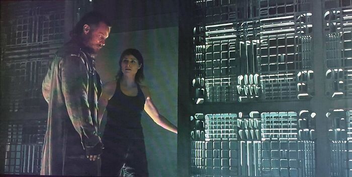 Two actors in a dimly lit set with creative wall props, one wearing a long coat and the other in a tank top, explore the scene.