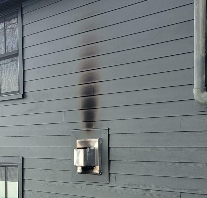 A vent on a gray exterior wall shows a dangerous black soot stain above it.
