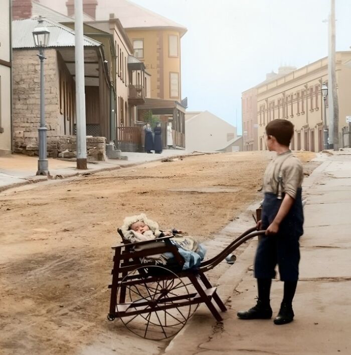 Boy pushes a baby in an old-fashioned stroller on a historical street, reflecting early 20th-century life.