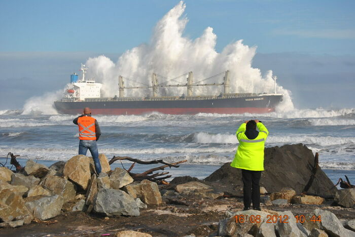 Two people photographing a massive wave crashing into a cargo ship, capturing a dramatic ocean scene from the rocky shore.