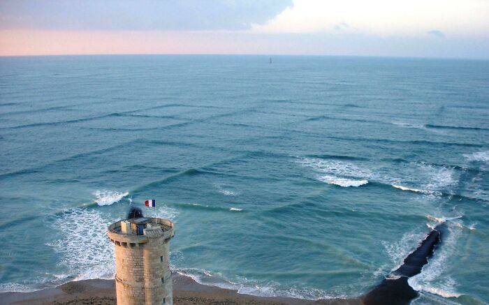 Cross sea waves near a coastal tower under a cloudy sky, creating an eerie ocean pattern.