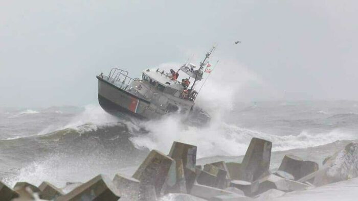A small boat struggling against large ocean waves during a storm near a rocky coast.