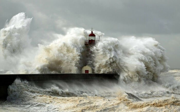 Lighthouse engulfed by massive ocean waves under a stormy sky.