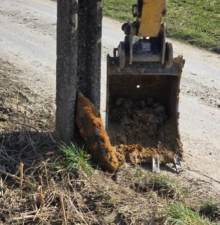 Excavator bucket digging dangerously close to a concrete pole on a dirt road.