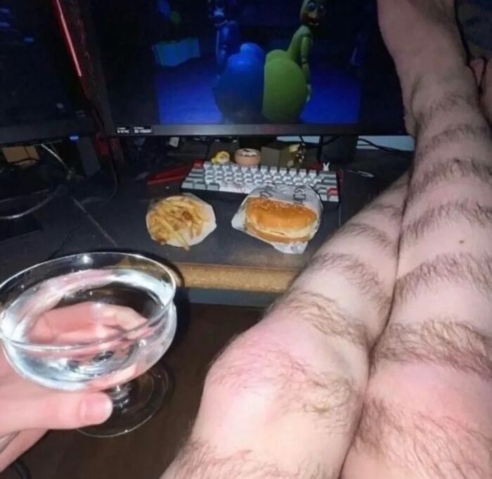 Person with hairy legs holding a drink, fast food on desk, gaming setup in background, representing random cursed images.