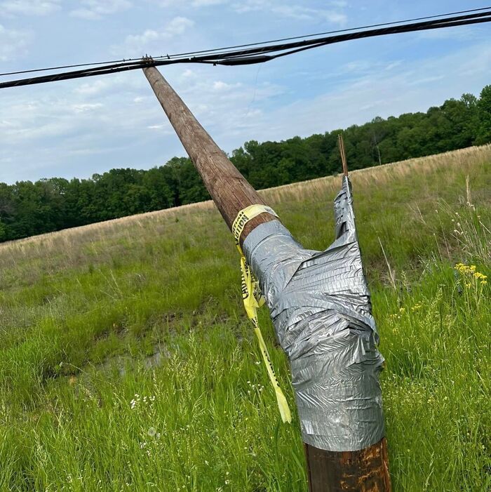 Failed repair attempt of a broken utility pole wrapped in duct tape and caution tape in a grassy field.