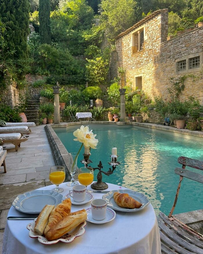 Table set by a picturesque pool, surrounded by lush greenery and a rustic stone building, showcasing a beautiful place.