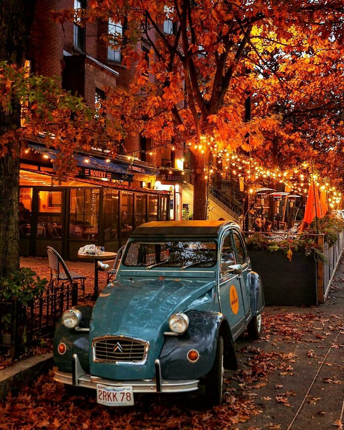 Vintage blue car on a leaf-covered road in front of a cozy café, surrounded by autumn trees and warm lights.