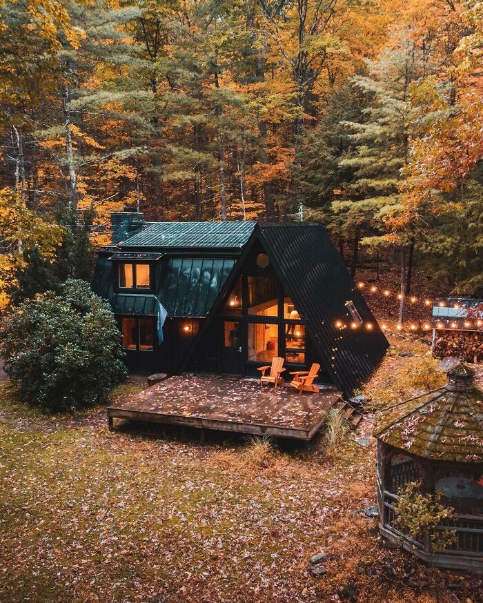 A-frame cabin surrounded by autumn forest, featuring string lights and a cozy wooden deck with chairs, in a beautiful place.