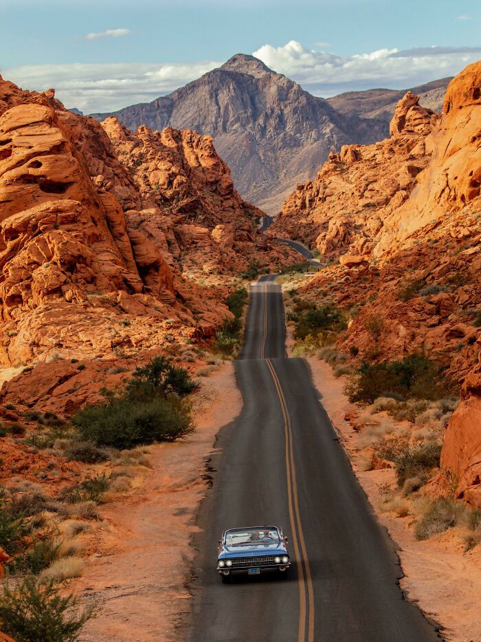 Vintage car on a scenic road through stunning red rock formations leading to a majestic mountain in the distance.