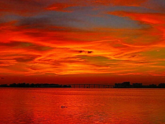 Stunning sunset over a tranquil lake with a silhouetted bridge and vibrant red clouds.