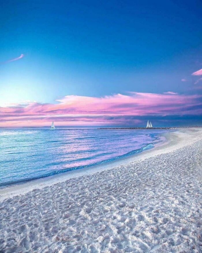 Sandy beach at sunset with pink clouds and sailboats on a tranquil sea, showcasing a beautiful place discovered.