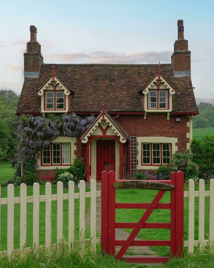 Charming brick cottage with a red door and lush garden behind a white picket fence, showcasing beautiful places.