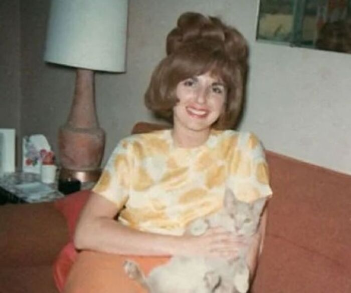 Woman with a vintage hairstyle from yesteryear, smiling on a couch with a cat.