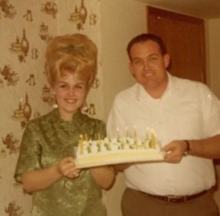 Vintage couple with a large beehive hairstyle, holding a cake with candles, embodying hairstyles from yesteryear.