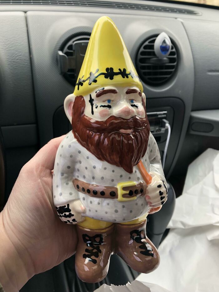 A hilarious gnome figurine with a yellow hat and beard, held inside a car, showcasing humorous style.