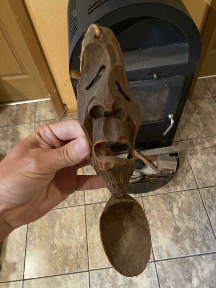 Mano sosteniendo cuchara tallada con rostro asustado frente a estufa y piso de baldosas en casa nueva.