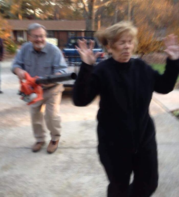 Elderly man playfully chases woman with leaf blower, showcasing humor.