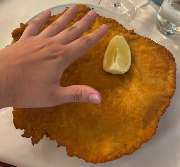 Hand comparing size to a large schnitzel, a renowned dish from different countries, with a lemon wedge on top.