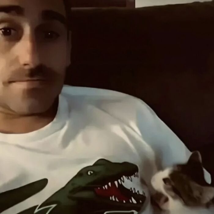 Man in a dinosaur shirt smiling at a cat on a couch; a funny random image from Instagram accounts.
