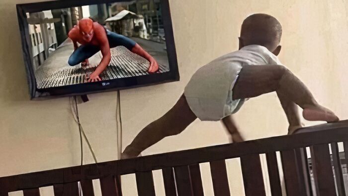 Baby mimicking Spider-Man on TV, climbing furniture humorously.