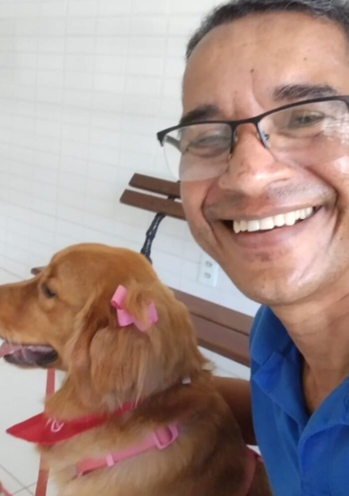 Man smiling with pet taxi passenger, a dog adorned with pink bows and bandana, sitting on a bench.