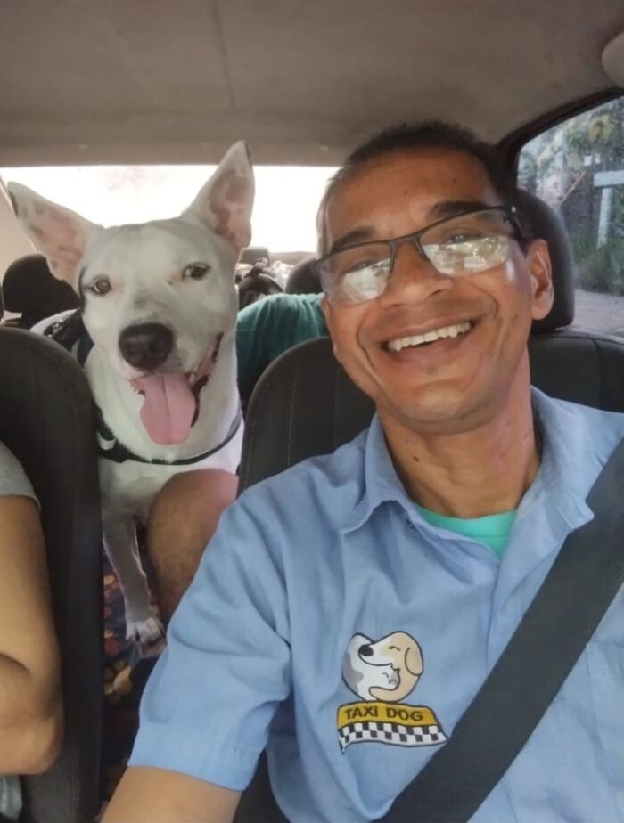 Man in glasses with smiling dog in a pet taxi, wearing a blue "TAXI DOG" shirt, capturing a selfie together.