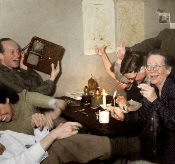 Group of people laughing around a table with a radio, evoking historical camaraderie and joy.
