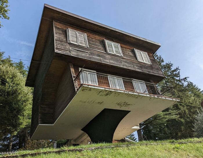 Upside-down house on inclined terrain, appearing fake but actually real, surrounded by trees under a clear sky.