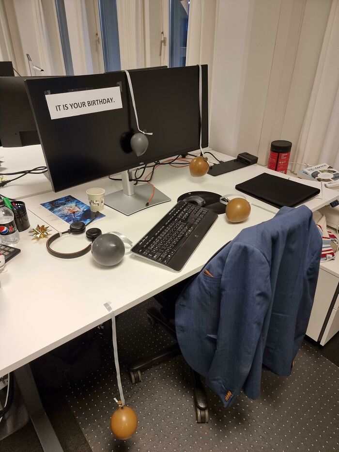 Office desk with funny birthday prank setup, featuring a sign reading "IT IS YOUR BIRTHDAY" and small balloons.
