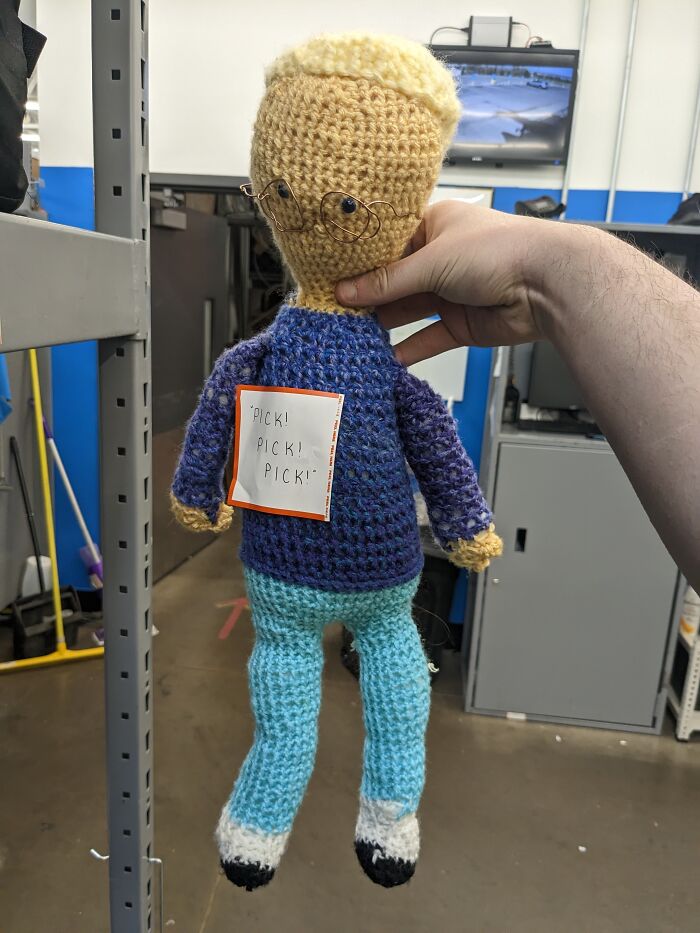 A knitted doll held in an office setting, showing a humor note reading “Pick! Pick! Pick!”.