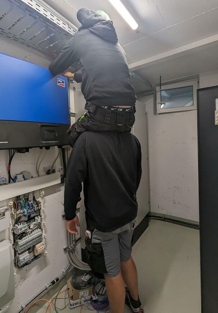 Two funny coworkers using one another for height to reach a high electrical panel in a utility room.
