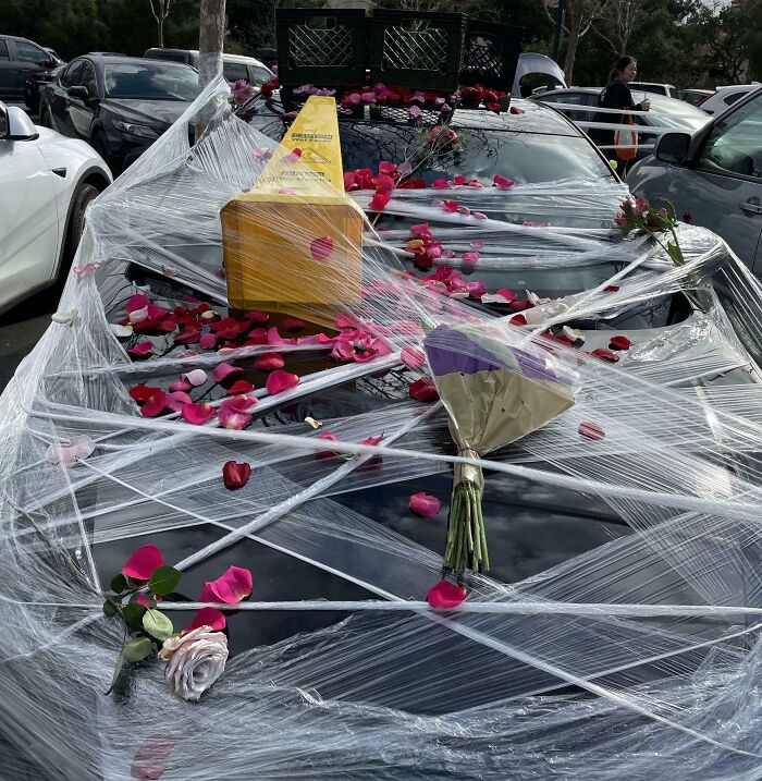 Car covered with plastic wrap, flowers, and petals as a funny coworker prank, showcasing office humor.
