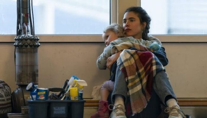 Woman hugging child, sitting on floor wrapped in a blanket, with cleaning supplies nearby, highlighting emotional movie scene.