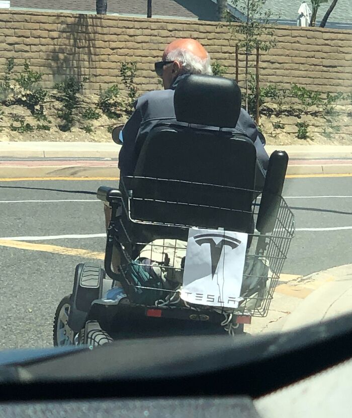 Elderly person humorously driving a scooter with a Tesla logo on the back.