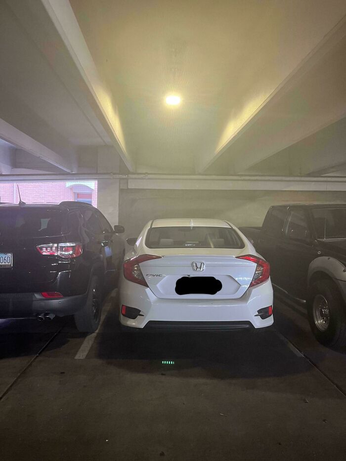 White car parked frustratingly between two larger vehicles in a dimly lit garage, highlighting online posting anger.