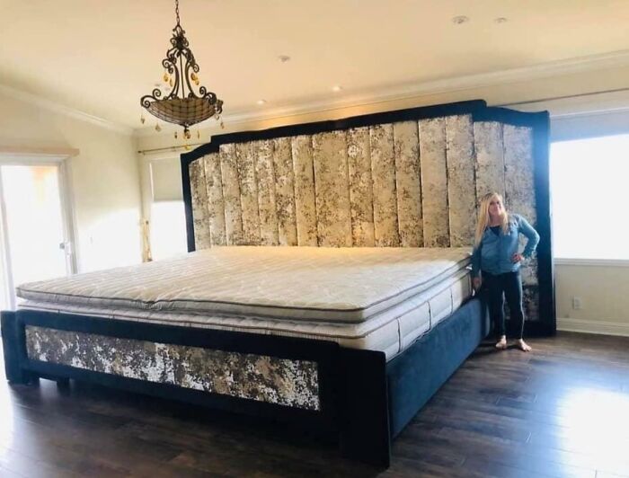 Giant bed appears fake due to its massive size, with a person standing beside for scale in a well-lit bedroom.