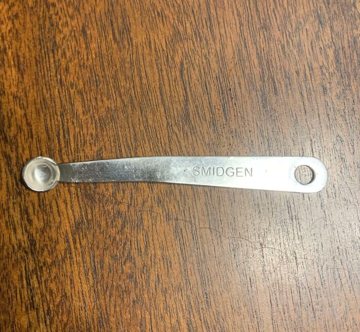Tiny metal measuring spoon labeled "Smidgen" on a wooden surface, looking fake but real.