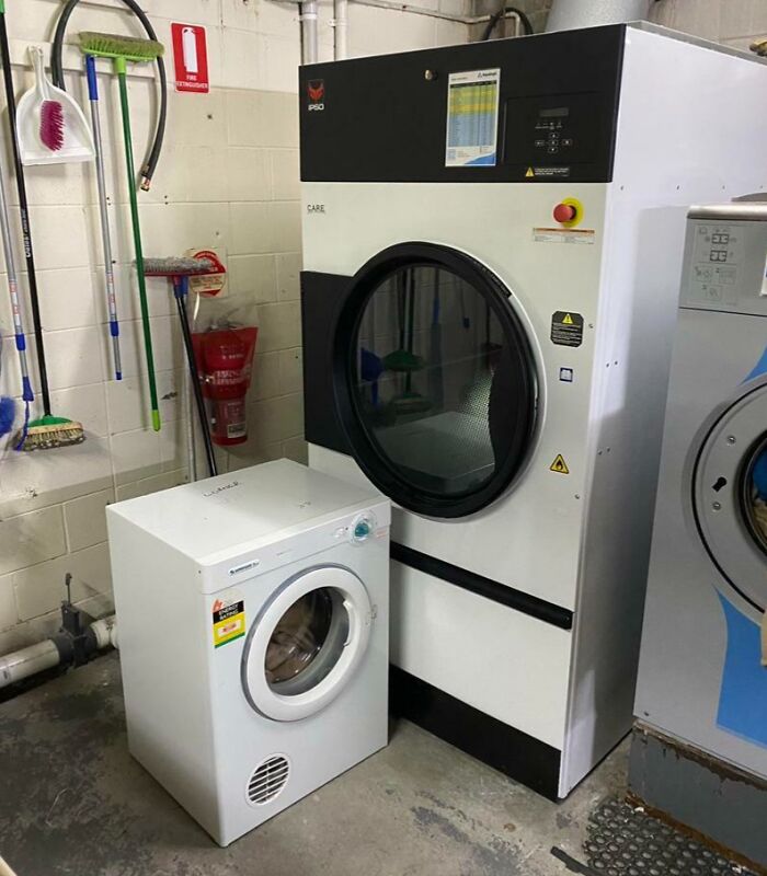 Small dryer next to an oversized industrial dryer in a laundry room, creating a surreal and fake-looking contrast.
