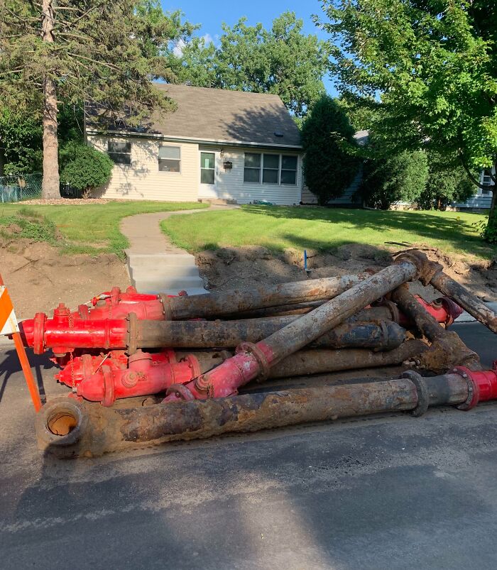 Pile of rusty red fire hydrants on a dug-up sidewalk, appearing unreal at first glance against a suburban house backdrop.